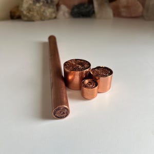 May include: A collection of four copper items consisting of one long cylindrical rod and three circular plugs of varying diameters. Each piece features a flat top surface with a detailed, intricate spiral pattern etched into the metallic copper.