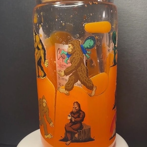Op de afbeelding: Een doorzichtige glazen beker met een bamboe deksel, gevuld met een oranje vloeistof en glitters. De buitenkant is voorzien van diverse illustraties van Bigfoot en een alien, waaronder een Bigfoot zittend op een boomstronk en een lopende Bigfoot.