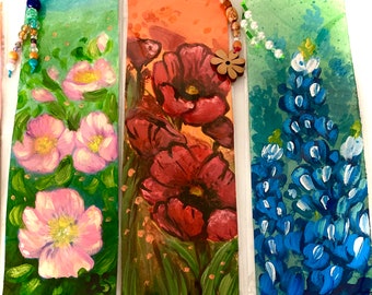 May include: Three hand-painted bookmarks featuring floral designs. The first bookmark has pink flowers on a green background. The second bookmark has red poppies on an orange background. The third bookmark has blue flowers on a green background.
