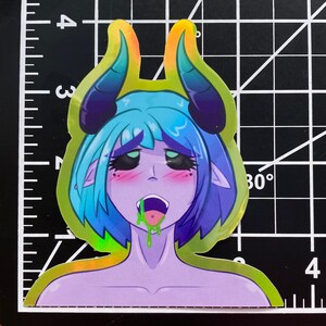 May include: A holographic sticker featuring a cartoon character with blue hair, purple skin, and green horns. The character has a large open mouth with green drool dripping from it. The sticker has a rainbow iridescent finish.