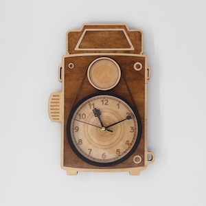 May include: A wooden wall clock shaped like a vintage camera. The clock face is circular with black numbers and hands. The clock is made of light and dark brown wood, with a detailed design mimicking a camera's features.