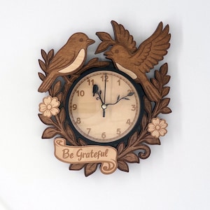 May include: Wooden wall clock featuring two birds, leaves, and flowers. The clock face is light-colored with black numbers and hands. The words "Be Grateful" are on a banner at the bottom. The clock is a decorative item.