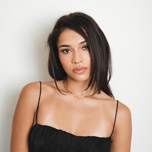 May include: A black spaghetti strap dress with a ruched bodice and a gold chain necklace. The dress is worn by a woman with dark hair. The woman is looking at the camera. The dress has a fitted silhouette.
