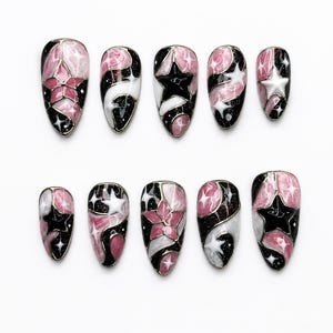 May include: A set of ten press-on nails featuring a celestial theme with a black, white, and pink marbled design. Each nail is decorated with star shapes, glitter accents, and thin silver lines that create a stained-glass effect across the surface.