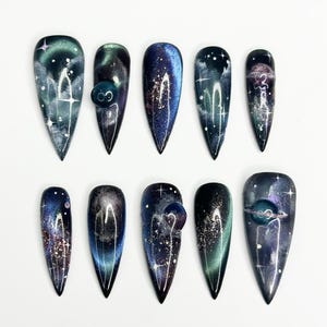 May include: A set of ten almond-shaped press-on nails featuring a dark galaxy theme with deep blue, purple, and black hues. The designs include shimmering star patterns, nebula-like swirls, and small raised spherical accents on select nails.