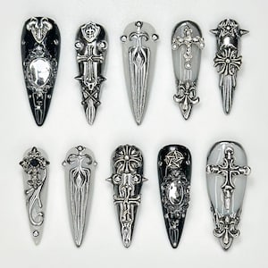 May include: A collection of ten gothic-style nail art designs. The nail art features silver-coloured embellishments with crosses, shields, and ornate patterns. Some nails are black, others are clear or light-coloured.