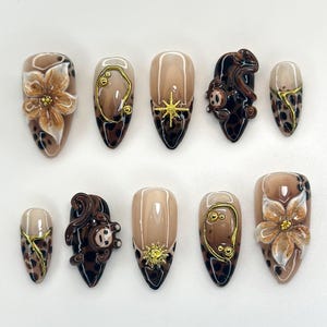 Könnte beinhalten: Ein Set aus zehn mandelförmigen Kunstnägeln. Die Nägel weisen eine Vielzahl von Designs auf, darunter florale Akzente, goldene Verzierungen und Tiermuster. Die Farbpalette umfasst Braun, Beige und Gold.