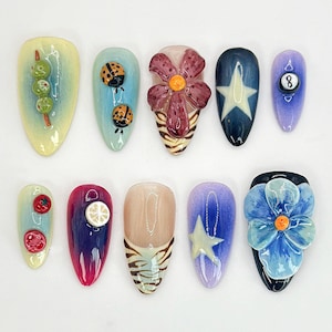 K&ouml;nnte beinhalten: Zehn verschiedene Nageldesigns mit verschiedenen 3D-Verzierungen. Die Designs beinhalten eine Blume, einen Stern, einen Marienk&auml;fer, eine Zitrone, eine Tomate, eine gr&uuml;ne Olive, einen Zebra-Druck und eine Billardkugel.