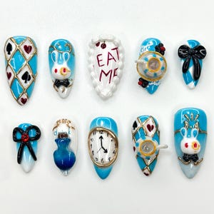 Puede incluir: Una colección de diez uñas artificiales con varios diseños. Las uñas presentan una base azul claro con detalles dorados e incluyen diseños como palos de cartas, un conejo blanco, una taza de té, un lazo, un reloj y el texto "EAT ME".