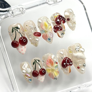 May include: A set of ten artificial nails with a clear base, adorned with cherries, flowers, mushrooms, and pearls. The nails feature a variety of colors, including red, white, and yellow, and are decorated with glitter and small gems. The nails are in a clear plastic case.
