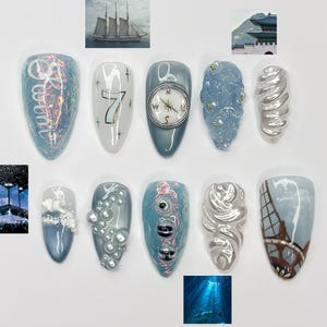 May include: A collection of ten artificial nails with various designs. The nails feature a range of colors, including blue, white, and silver, with embellishments like a compass, the number 7, and a depiction of the Eiffel Tower. Some nails have textured surfaces.