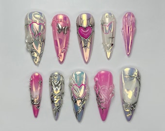 May include: A collection of ten long, pointed artificial nails. Each nail features a glossy, iridescent finish with pink, silver, and purple hues. Some nails have heart, butterfly, and floral designs, with intricate silver embellishments.