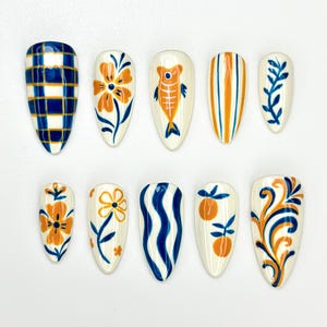 Puede incluir: Un conjunto de diez uñas postizas en forma de almendra con varios diseños pintados a mano. Los diseños incluyen cuadros, flores, peces, rayas y espirales en tonos azules, naranjas y blancos. Las uñas están dispuestas sobre una superficie blanca.
