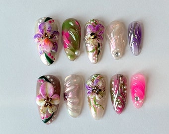 May include: Set of ten false nails with a floral design. The nails are painted with a white base and feature pink, purple, and green flowers. Some nails have a pearl or rhinestone accent.
