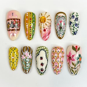 May include: A collection of ten painted artificial nails. Designs include a sun, flowers, and geometric patterns in various colors like pink, yellow, green, and blue. Each nail is a different design, showcasing a variety of artistic styles.