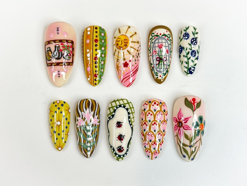 May include: A collection of ten painted artificial nails. Designs include a sun, flowers, and geometric patterns in various colors like pink, yellow, green, and blue. Each nail is a different design, showcasing a variety of artistic styles.