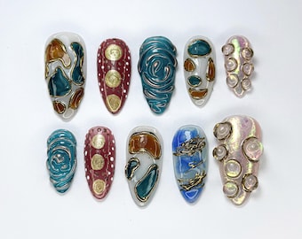 Puede incluir: Diez diseños de uñas diferentes que presentan patrones abstractos en tonos azules, marrones y dorados. Los diseños incluyen remolinos, puntos y formas geométricas.