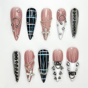 May include: A set of ten long, almond-shaped press-on nails. The nails feature a variety of designs, including solid colors, plaid patterns, and embellishments like chains, crosses, and studs. Colors include pink, white, black, and silver.