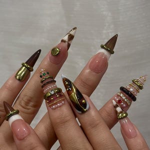 May include: A set of long, pointed artificial nails. The nails feature a variety of designs, including brown and white checkered patterns, gold accents, and decorative rings with pearls and gems. The nails have a natural pink base with white tips.