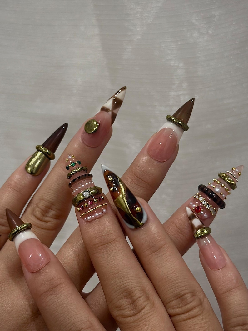 May include: A set of long, pointed artificial nails. The nails feature a variety of designs, including brown and white checkered patterns, gold accents, and decorative rings with pearls and gems. The nails have a natural pink base with white tips.