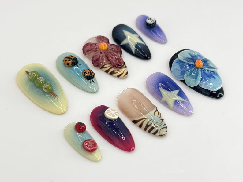 K&ouml;nnte beinhalten: Zehn verschiedene Nageldesigns mit verschiedenen Themen, darunter Blumen, Marienk&auml;fer, Sterne und Fr&uuml;chte. Die Designs sind auf wei&szlig;em Hintergrund gemalt.