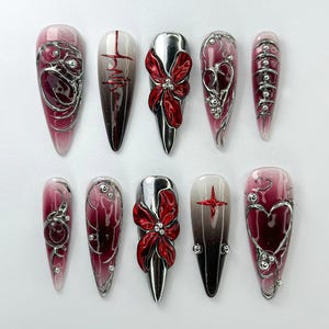 May include: Ten long, almond-shaped false nails with different designs. Each nail is painted with a gradient of white and red, and features a silver 3D design. The designs include a heart, a flower, and a swirl.