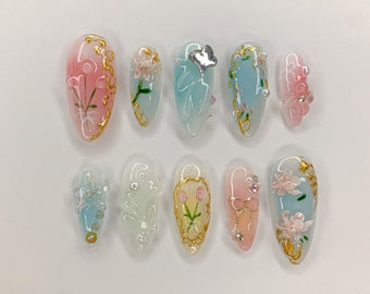 May include: Ten oval-shaped false nails with a light blue, pink, or white base. Each nail is decorated with gold accents, flowers, and glitter.