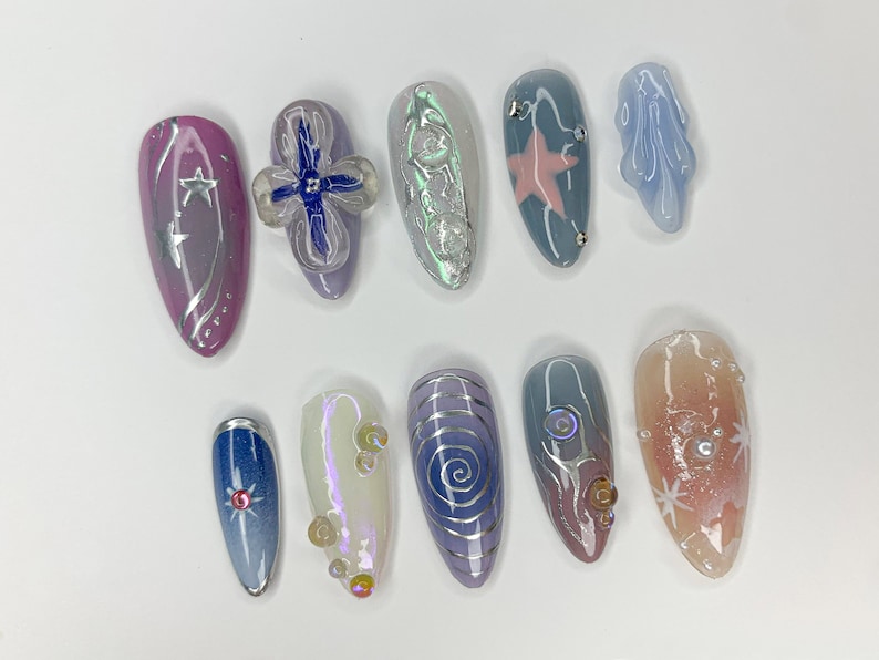 May include: A set of ten long, oval-shaped artificial nails with various designs. The nails are painted in shades of blue, pink, and white with silver accents. Some nails feature stars, swirls, and a flower design.