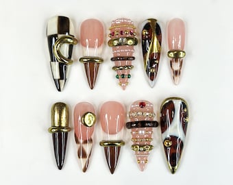 May include: A collection of ten artificial nails in various designs. Some nails feature a checkerboard pattern, while others have a nude base with gold or brown accents. Several nails are adorned with rings and jewels.