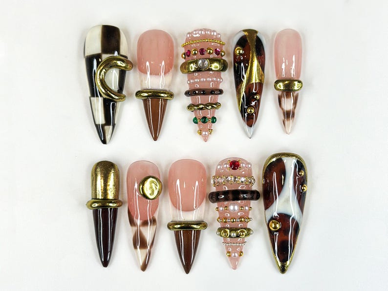 May include: A collection of ten artificial nails in various designs. Some nails feature a checkerboard pattern, while others have a nude base with gold or brown accents. Several nails are adorned with rings and jewels.