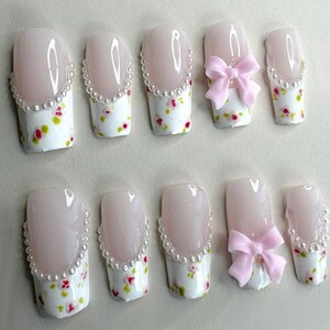 Può includere: Set di dieci unghie finte. Ogni unghia presenta una base rosa chiaro, un motivo floreale bianco e un bordo di perle. Alcune unghie hanno un fiocco rosa. Progettate per la manicure.