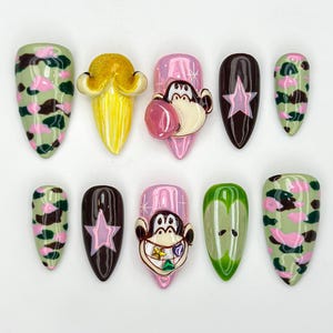 May include: A collection of ten almond-shaped false nails. Designs include a yellow sculpted nail, pink nails with a monkey face, brown nails with a pink star, and green camouflage nails with pink and brown accents.