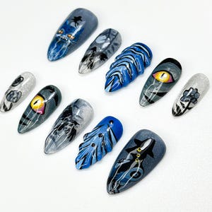 May include: A set of ten long, almond-shaped artificial nails with various designs. The nails feature detailed artwork, including a blue monster, an eye, and abstract patterns in shades of blue, gray, and silver. The nails are arranged on a white surface.