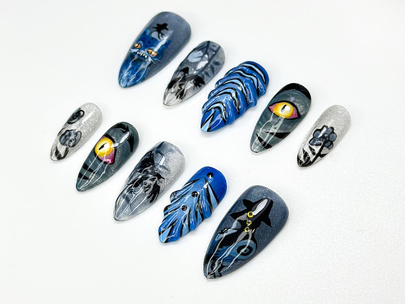 May include: A set of ten long, almond-shaped artificial nails with various designs. The nails feature detailed artwork, including a blue monster, an eye, and abstract patterns in shades of blue, gray, and silver. The nails are arranged on a white surface.