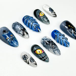 May include: A collection of ten painted artificial nails. Designs include a blue cat, an eye, and abstract patterns in shades of blue, gray, and white. The nails are arranged on a white surface.