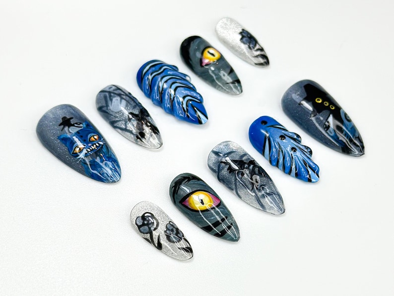 May include: A collection of ten painted artificial nails. Designs include a blue cat, an eye, and abstract patterns in shades of blue, gray, and white. The nails are arranged on a white surface.