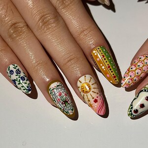 May include: A hand with long, almond-shaped nails, each uniquely decorated. Designs include floral patterns, a sun, and geometric shapes in various colors like green, pink, and white. The nails are painted with intricate details.