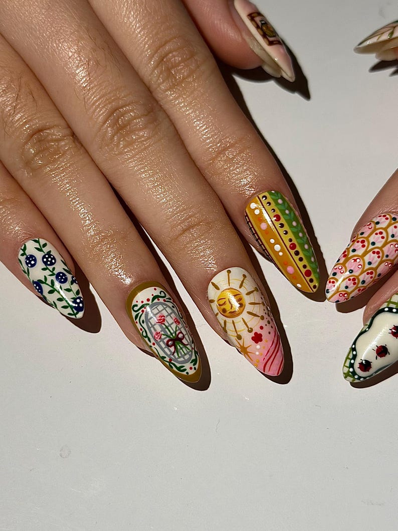 May include: A hand with long, almond-shaped nails, each uniquely decorated. Designs include floral patterns, a sun, and geometric shapes in various colors like green, pink, and white. The nails are painted with intricate details.