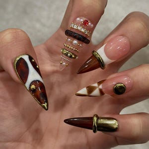 May include: A hand displaying a set of long, pointed acrylic nails. The nails feature various designs, including brown and white patterns, gold accents, and embedded gems. Some nails have a French tip design with a brown cone shape.