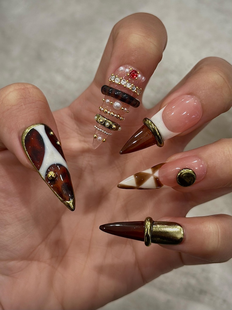 May include: A hand displaying a set of long, pointed acrylic nails. The nails feature various designs, including brown and white patterns, gold accents, and embedded gems. Some nails have a French tip design with a brown cone shape.