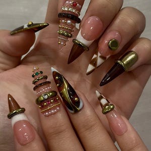 May include: A set of long, pointed acrylic nails with various designs. Some nails feature a nude base with white tips and brown accents. Others have intricate embellishments, including gold rings, rhinestones, and pearl-like beads. The overall aesthetic is glamorous and detailed.