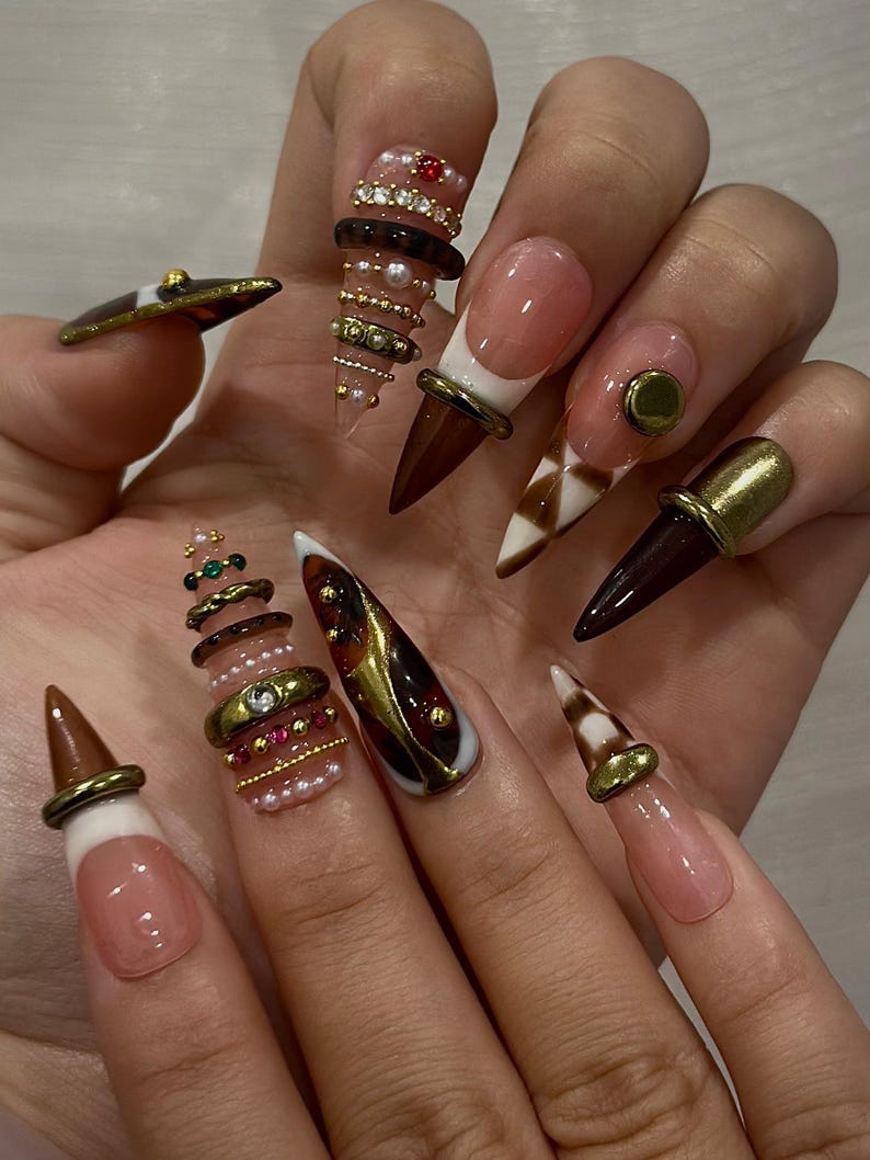 May include: A set of long, pointed acrylic nails with various designs. Some nails feature a nude base with white tips and brown accents. Others have intricate embellishments, including gold rings, rhinestones, and pearl-like beads. The overall aesthetic is glamorous and detailed.