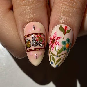 May include: Close-up of two fingernails with detailed nail art. One nail features a design with jars, a vase, and a skull. The other nail showcases a floral design with pink and blue flowers and green leaves. The nails are painted on a light pink base.