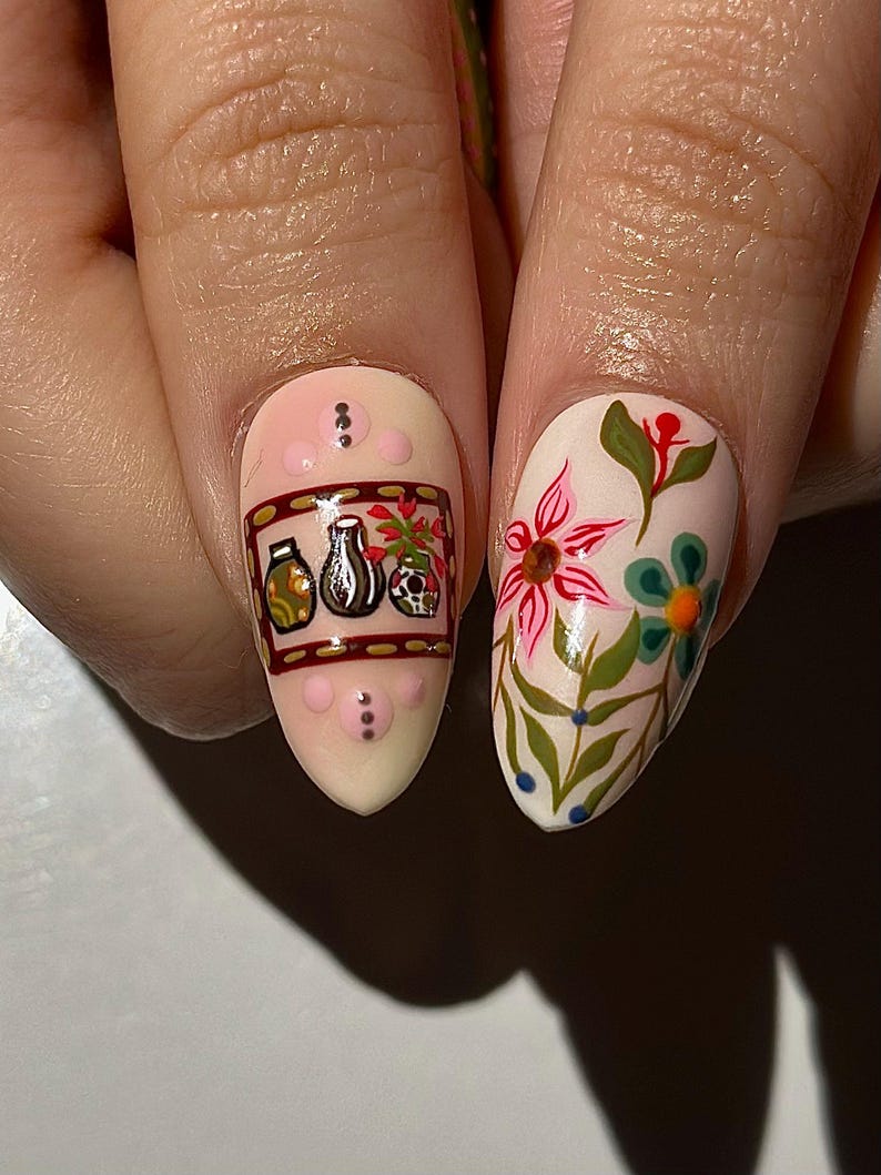 May include: Close-up of two fingernails with detailed nail art. One nail features a design with jars, a vase, and a skull. The other nail showcases a floral design with pink and blue flowers and green leaves. The nails are painted on a light pink base.