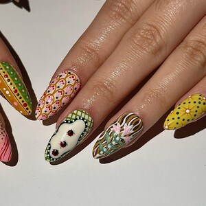 May include: Close-up of a hand with long, painted fingernails. The nails feature various designs, including polka dots, floral patterns, and geometric shapes in colors like pink, green, yellow, and brown. The nails are almond-shaped.