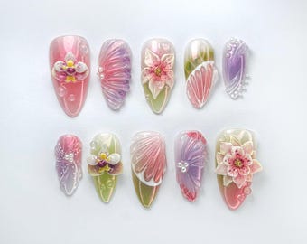 Puede incluir: Diez uñas postizas con diferentes diseños florales. Las uñas están pintadas en tonos de rosa, morado y verde con purpurina iridiscente. Algunas de las flores son 3D y tienen detalles de perlas.