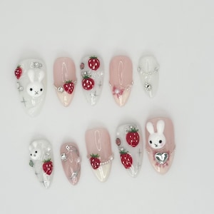 Pode incluir: Conjunto de dez unhas artificiais rosa e brancas com tema de morango e coelho. As unhas são decoradas com pequenos strass e pingentes 3D de coelho e morango.