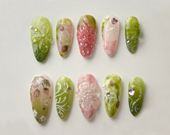 May include: Ten green and white almond-shaped artificial nails with various floral designs, including strawberries, flowers, and vines. Some nails have small rhinestones.