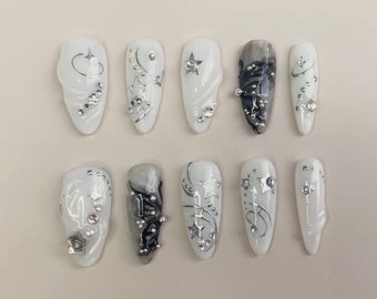May include: Ten white and gray long oval-shaped artificial nails with silver and black designs. The nails have a swirl design with stars and rhinestones.