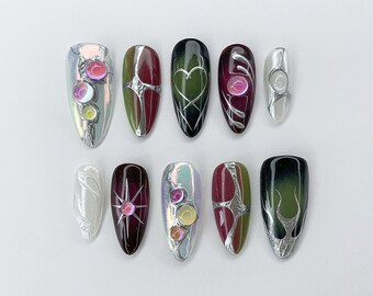 May include: Ten different nail designs featuring a variety of colors and patterns, including green, red, black, and white. Some nails have iridescent accents and silver foil designs.